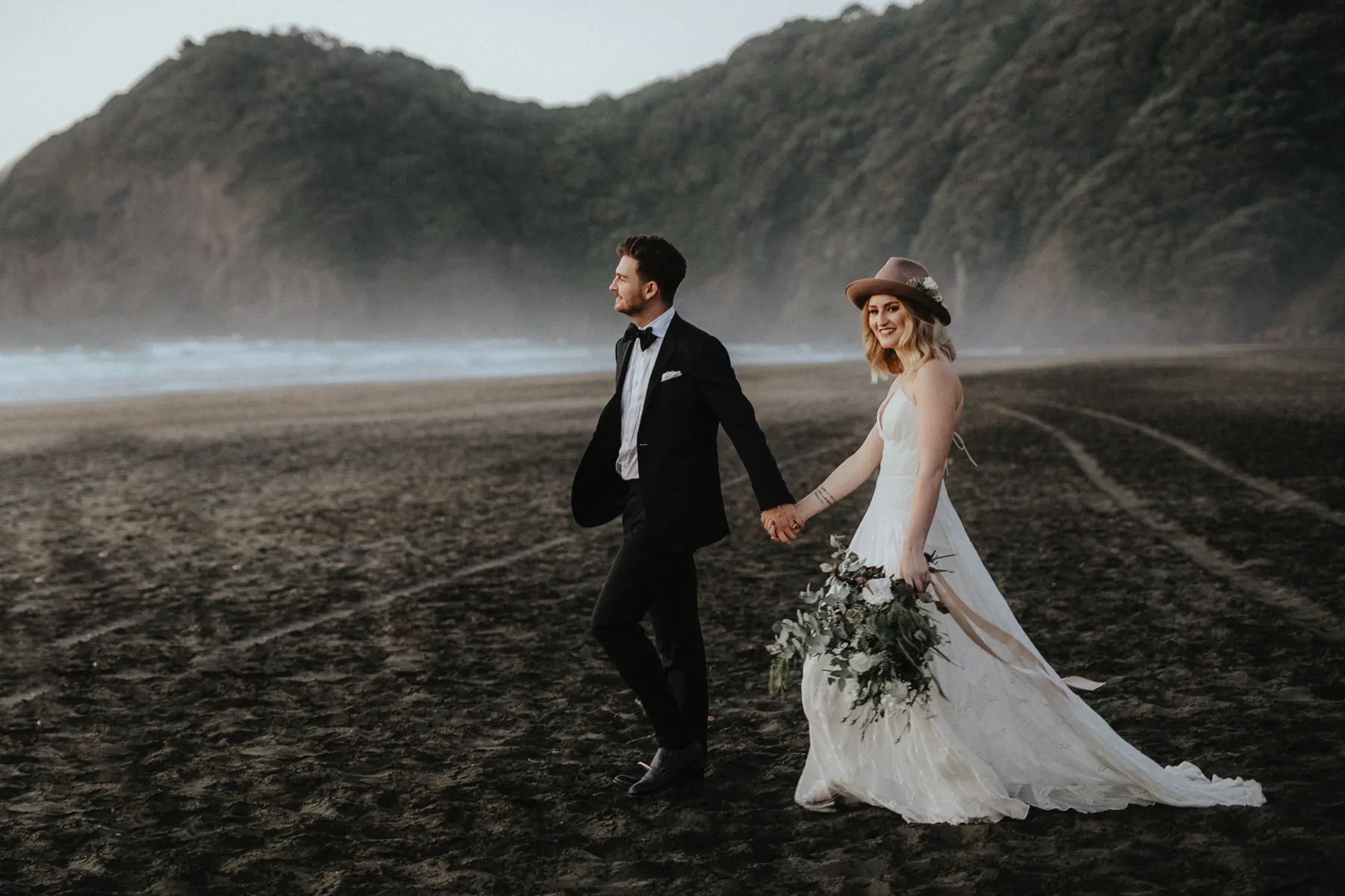 A bride and groom walking hand in hand along a misty black-sand beach